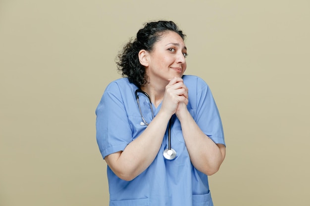 Begging middleaged female doctor wearing uniform and stethoscope around neck looking at camera keeping hands together on chin showing please gesture isolated on olive background