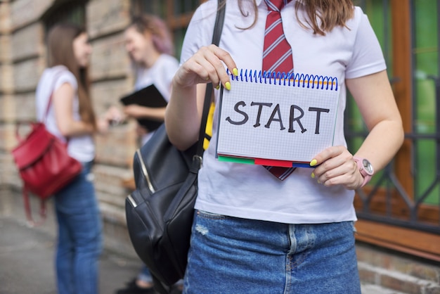 Begin. Meisje student houdt notitieboekje met woord start, begin van de lessen op school, op de universiteit. Bakstenen gebouw en pratende studenten achtergrond