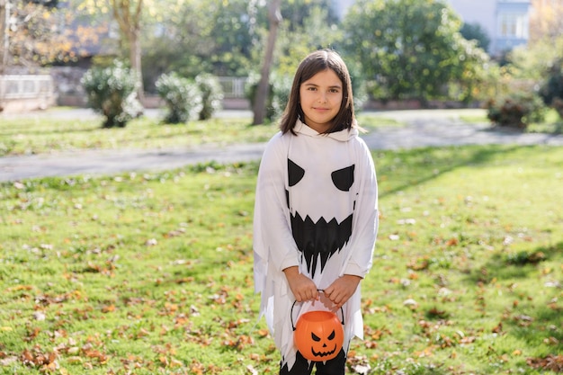 Foto behandelen of bedriegen buitenopname van lachend meisje in spookcarnaval-outfit voor halloween die in het park staat met pompoenmand in haar handen ruimte kopiëren