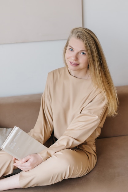 Beige cozy interior and a beautiful young woman in a stylish beige suit with a book. Concepts of home and comfort.