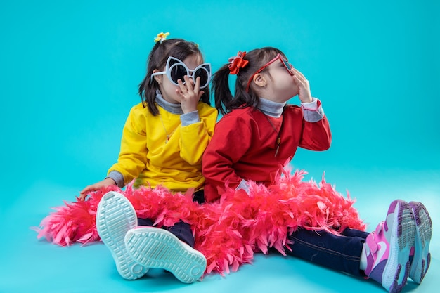 Being carefree together. Two happy girls sitting on studio floor while wearing glasses and covering legs with red festive boa