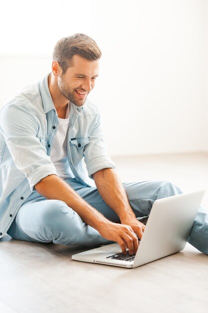 Being at home in pleasure. Handsome young man working on laptop and smiling while sitting on the floor at his apartment