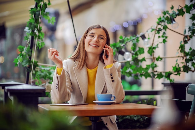 Beïnvloeder zittend op terras van cafetaria, koffiepauze en telefoneren met vriend.