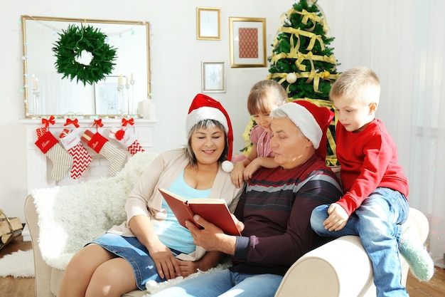 Bejaard echtpaar leest boek aan hun kleinkinderen in woonkamer ingericht voor Kerstmis decorated