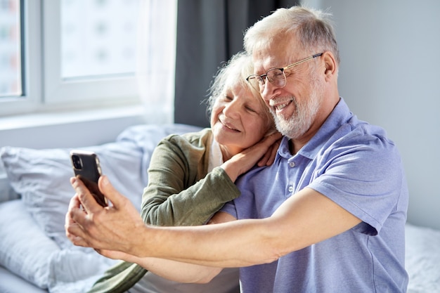 bejaarde echtpaar nemen foto op smartphone, zittend in de slaapkamer, zitten glimlachen. senior mensen samenleving levensstijl technologie concept. man en vrouw delen sociale media samen in welzijnshuis