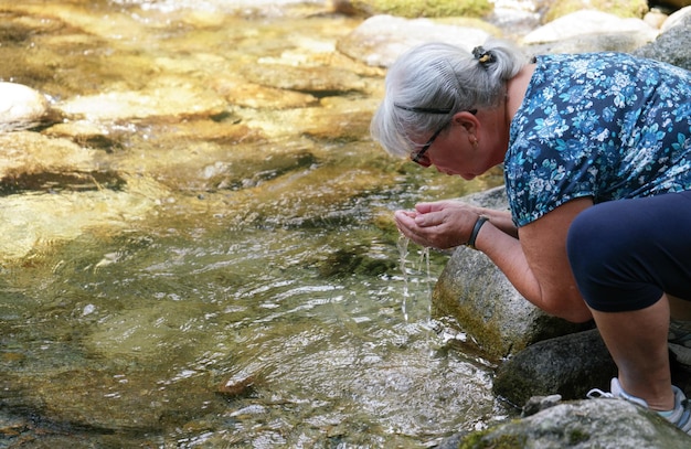 Bejaarde oudere vrouw die water uit de bosrivier vasthoudt in een kopje van haar handen die op het punt staat het te drinken