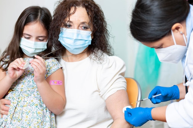 Bejaarde vrouw die een gezichtsmasker draagt met haar kleindochter die een dosis Coronavirus-vaccin krijgt. Vaccinatieconcept bij patiënten met een hoog infectierisico.