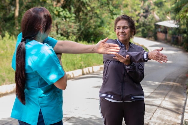 Bejaarde vrouw gekleed in sportkleding glimlachend terwijl ze schouderrekoefeningen uitvoert onder begeleiding van een verpleegster
