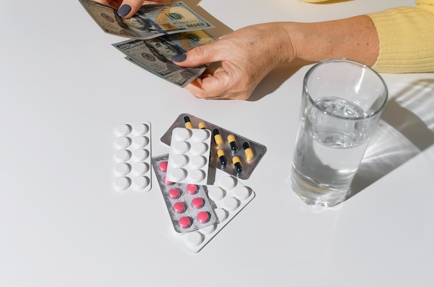 Bejaarde vrouw handen met geld. Tabletten en glas water op witte tafelblad weergave. Concept behandelingskosten, medicijnprijs, ziekteverzekeringsbudget