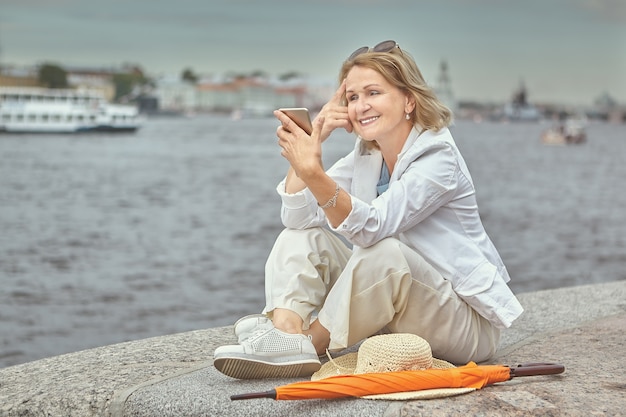 Bejaarde witte vrij lachende dame 62 jaar oud zit bij de rivier tijdens een wandeling in de binnenstad met mobiele telefoon in handen.