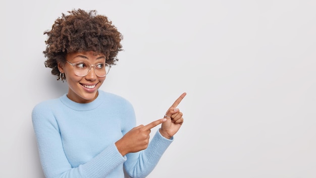 Bekijk dit eens. positieve mooie vrouw met krullend haar glimlacht gelukkig geeft op lege ruimte gebied toont plaats voor uw advertentie draagt bril blauwe trui geïsoleerd op witte achtergrond