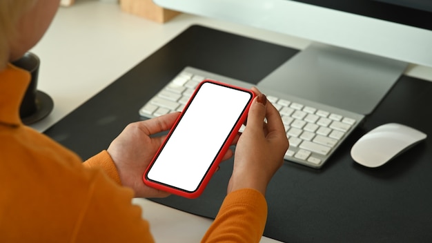 Bekijk over schouder vrouw met mobiele telefoon met wit scherm zittend aan haar bureau.