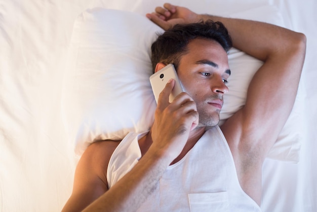 Bekijk van bovenaf het portret van een knappe jongeman die een mobiele telefoon in het witte bed gebruikt