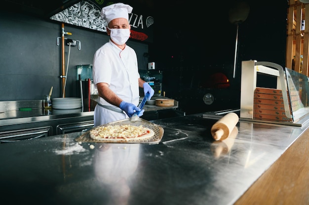 Bekwame chef-kok die traditionele Italiaanse pizza bereidt in het interieur van de moderne restaurantkeuken met speciale houtgestookte oven. Het dragen van een beschermend medisch gezichtsmasker en handschoenen in het nieuwe normale concept van het coronavirus