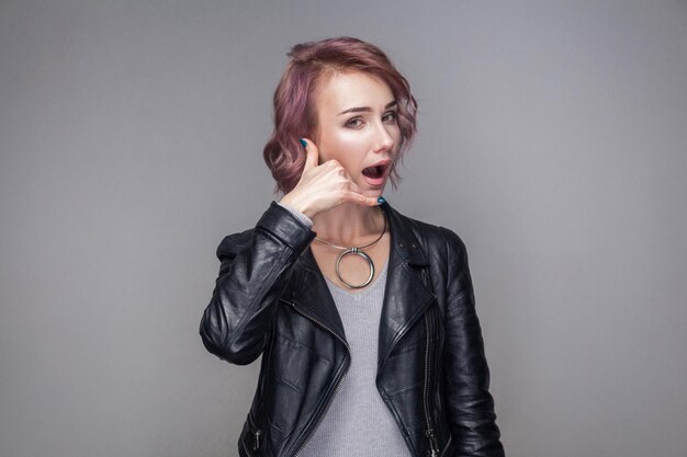 Bel mij. Portret van een grappig mooi meisje met kort haar en make-up in een zwarte leren jas in een casual stijl, die naar de camera kijkt met een oproepgebaar. indoor studio-opname, geïsoleerd op een grijze achtergrond