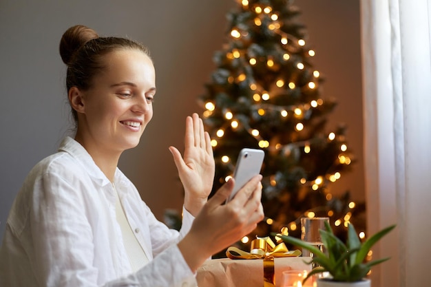 Bel vrienden en familie voor kerstwensen Vrouw met smartphone die geniet van een mobiel telefoongesprek terwijl ze in de buurt van de kerstboom zit en iedereen vrolijk kerstfeest wenst zwaaiend hallo gebaar