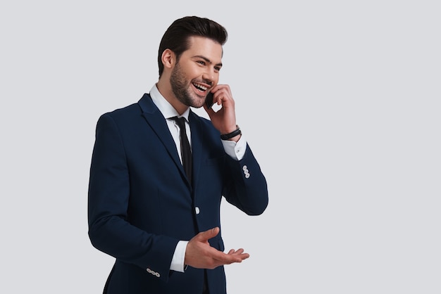 Belangrijk gesprek. Knappe jongeman in volledig pak die op zijn smartphone praat en glimlacht terwijl hij tegen een grijze achtergrond staat