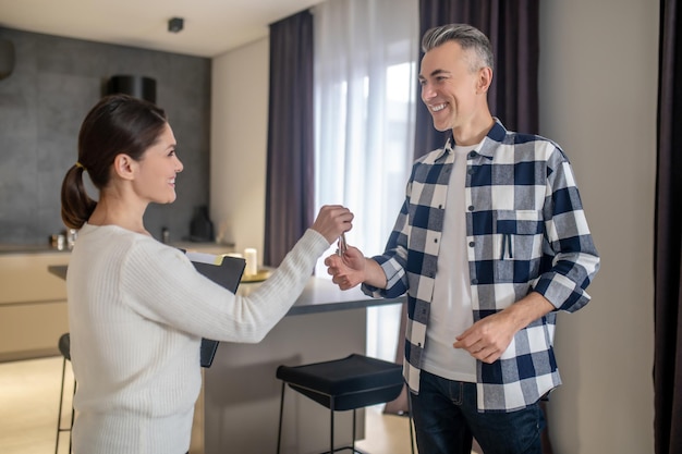 Foto belangrijk punt donkerharige lachende vrouw met map die sleutels geeft aan een gelukkige man van middelbare leeftijd