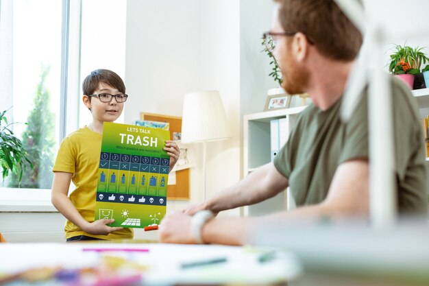 Belangrijke zaken. Geïnteresseerd donkerharig jongetje in heldere glazen met poster met instructies over afvalrecycling