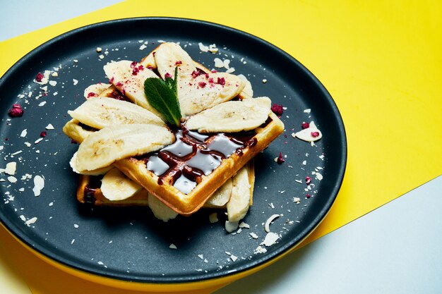 Belgian waffles with chocolate, bananas and almonds on a black plate on a colored surface Appetizing sweet pastries. Breakfast food