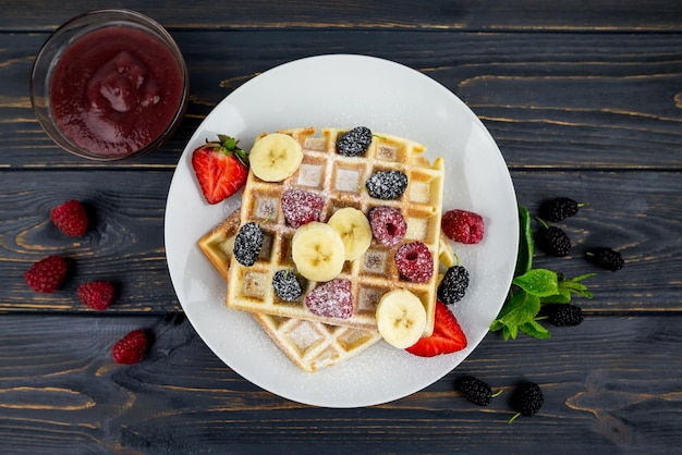 Belgian waffles with summer berries and powdered sugar in a white plate on a dark wooden background Sweet Belgian waffles for breakfast or lunch