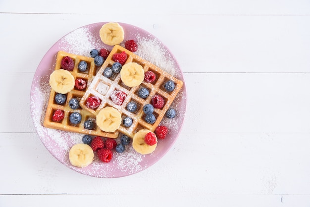 Belgische wafels met bosbessen, frambozen, bananen en suikerpoeder op witte achtergrond. Concept van lekker en gezond eten. Bovenaanzicht.