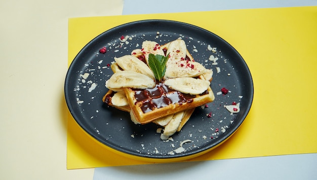 Belgische wafels met chocolade, bananen en amandelen op een zwarte plaat op een gekleurde ondergrond. Smakelijke zoete gebakjes. Ontbijt