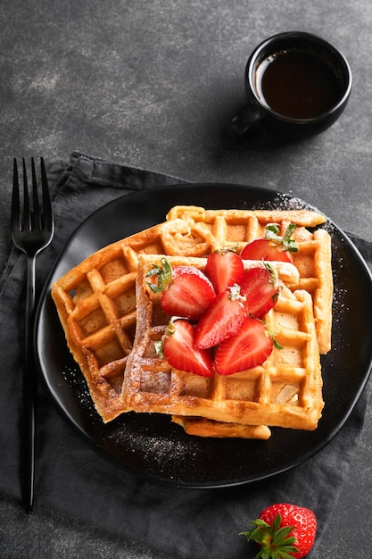 Belgium waffles Homemade waffles with strawberries powdered sugar and cup of coffee on black plate on black stone table background Breakfast Top view Mockup for design idea