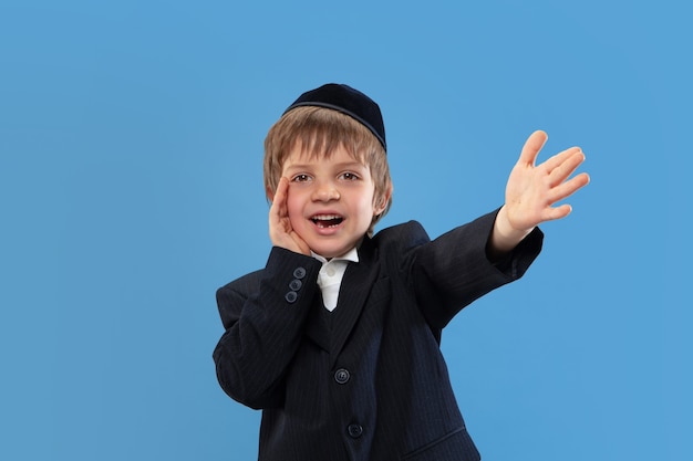 Bellen, schreeuwen. Portret van een jonge orthodoxe joodse jongen geïsoleerd op blauwe muur.