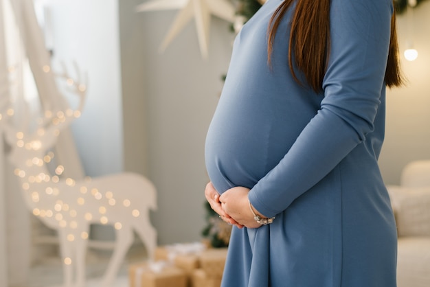 Belly of a pregnant woman in a knitted blue dress who holds her hands in the lower abdomen against the background of a Christmas glowing deer