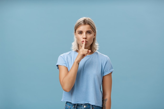 Beloof het niemand het geheim te vertellen. Mysterieuze aantrekkelijke en vrouwelijke blonde vrouw met een gebruinde huid en tatoeages die met wijsvinger over de mond dichtknijpen alsof ze iets over de blauwe muur verbergt