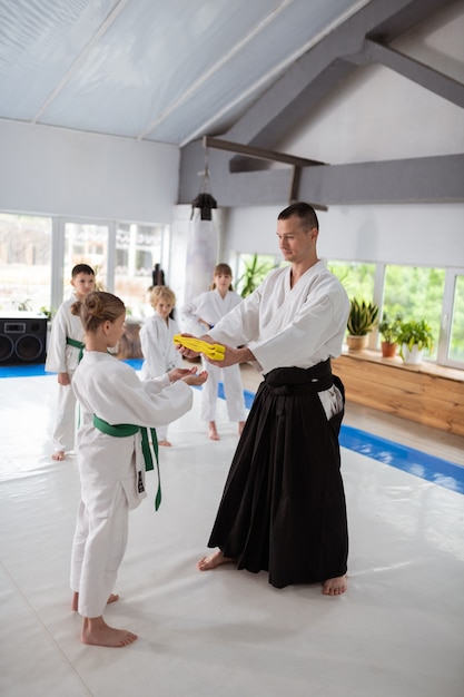 Belt for pupil. Young aikido trainer giving yellow belt to his beautiful talented pupil