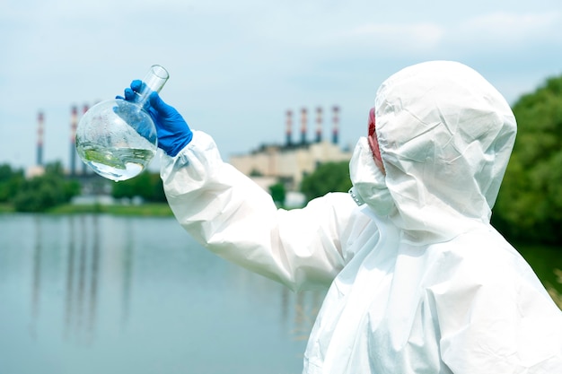 Bemonstering uit open water. een wetenschapper of bioloog neemt een watermonster in de buurt van een fabrieksinstallatie. een monster van water in een ronde chemische kolf.