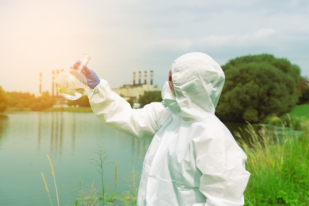 Bemonstering uit open water. Een wetenschapper of bioloog neemt een watermonster in de buurt van een fabrieksinstallatie. Een monster van water in een ronde chemische kolf.