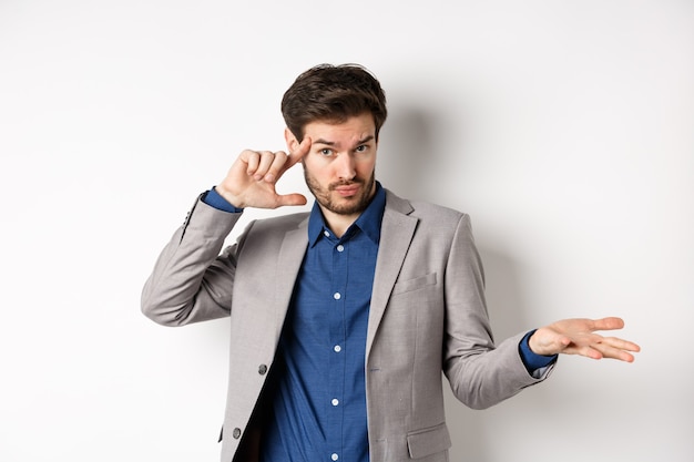 Ben je gek. jonge man in pak wijzende vinger op het hoofd om vreemde of domme fout uit te schelden, schouders ophalend verward, staande op een witte achtergrond.