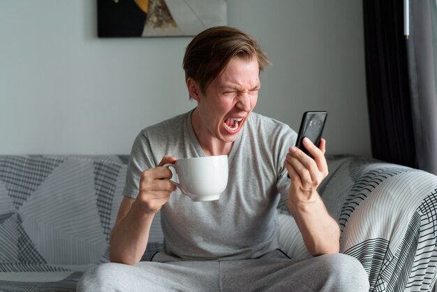 Benadrukt jonge man koffie te houden terwijl hij thuis aan de telefoon schreeuwde
