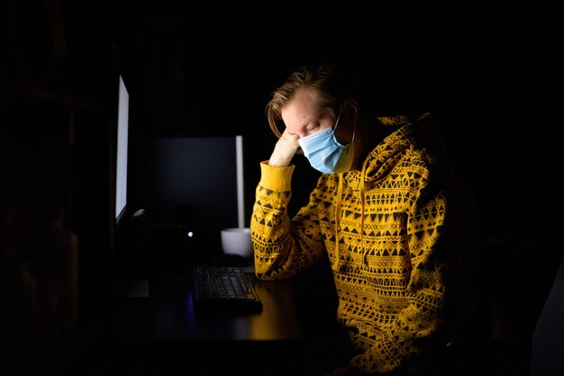 Benadrukt jongeman met masker op zoek moe tijdens het werken vanuit huis 's avonds laat in het donker