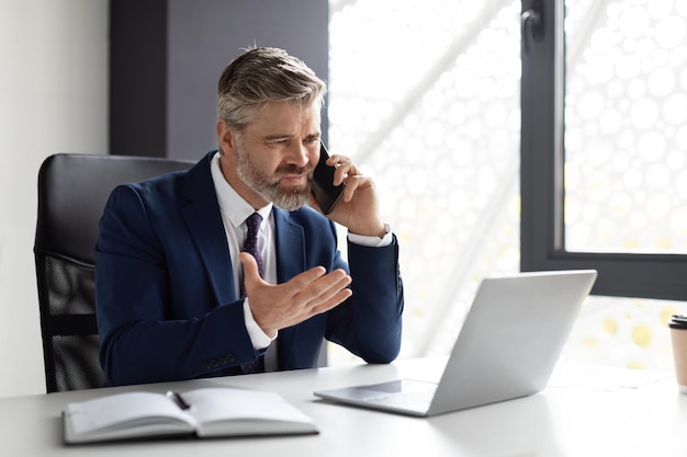 Benadrukt zakenman van middelbare leeftijd praten op mobiele telefoon en met behulp van laptop in office