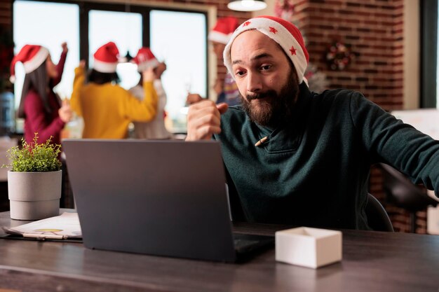 Benadrukte overbelaste blanke man in kerstmuts die op laptop werkt tijdens de kerstperiode. Uitgeputte werknemer die de projectdeadline beheert op de computer op kantoor tijdens de nieuwjaarsvakantie