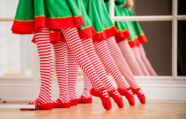 Benen van kinderen gekleed in kostuums van kerstelfen die oefenen in een balletbar voor een spiegel in een ruime witte studio.