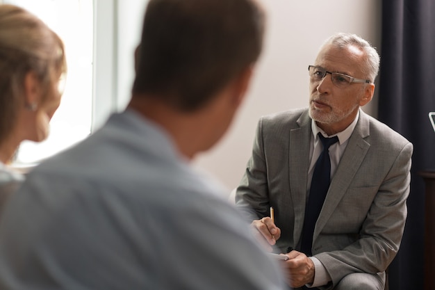 Beoordeling van de geestelijke gezondheid. Ernstige geconcentreerde psychoanalyticus met een bril die in zijn kantoor voor zijn patiënten zit terwijl hij hun geestelijke gezondheid beoordeelt