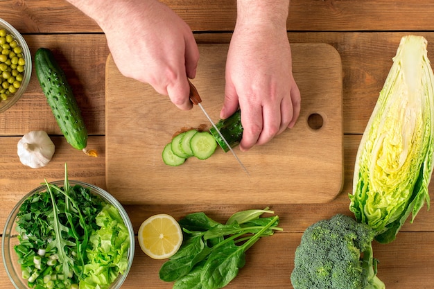 Bereiding van salade van verse groenten. Mannenhanden snijden de komkommer met een mes op een snijplank.