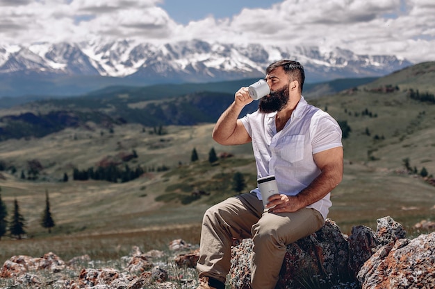 Berglandschap en groene vallei. Volwassen man met baard in een wit overhemd zit op een geplaveide steen en drinkt rea uit een mok en houdt een thermoskan vast om te reizen. Kijk opzij naar het prachtige uitzicht.