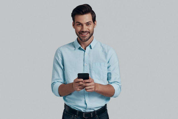 Bericht aan het typen. Knappe jonge man die slimme telefoon gebruikt en naar de camera kijkt terwijl hij tegen een grijze achtergrond staat