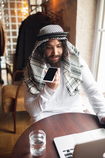 Bericht. arabische zakenman die op kantoor werkt, zakencentrum met apparaat, gadget. moderne saoedische levensstijl. man in traditionele kleding en sjaal ziet er zelfverzekerd, druk, knap uit. etniciteit, financiën.