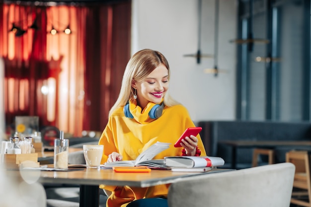 Bericht lezen. Vrouw met blauwe oortelefoons en gele trui die een bericht op de telefoon leest