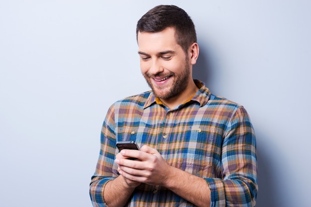 Bericht met mooie woorden. knappe jonge man in shirt die mobiele telefoon vasthoudt en ernaar kijkt terwijl hij tegen een grijze achtergrond staat