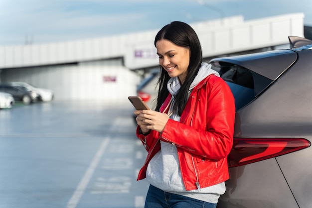 Bericht versturen. Vrolijk meisje leunt op haar nieuwe auto en typt op haar smartphone met een brede glimlach terwijl ze op de vlucht wacht. Mensen en gadgets concept