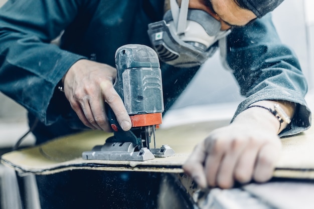 Beroep, mensen, timmerwerk, emotie en mensenconcept - een timmerman snijdt een bord met een elektrische puzzel. vervaardiging van skateboards. hij heeft beschermende kleding en een masker. 4k