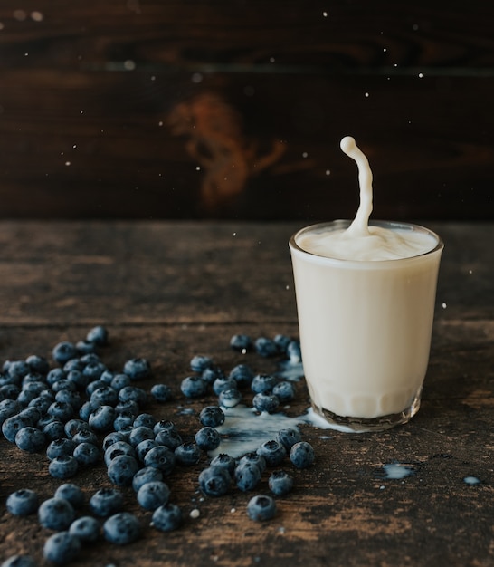 Berries are thrown into the transparent glass making splashes of milk. Blue fresh blueberries are scattered on an old brown wooden cracked table. White water pour down on the table.